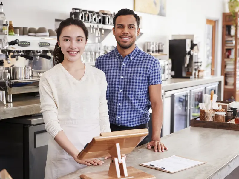 A business directory–style portrait of two smiling café owners standing behind the service counter in their coffee shop